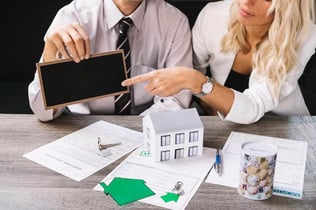Man and woman discussing home loan or financial documents with house model and blank chalkboard on table.